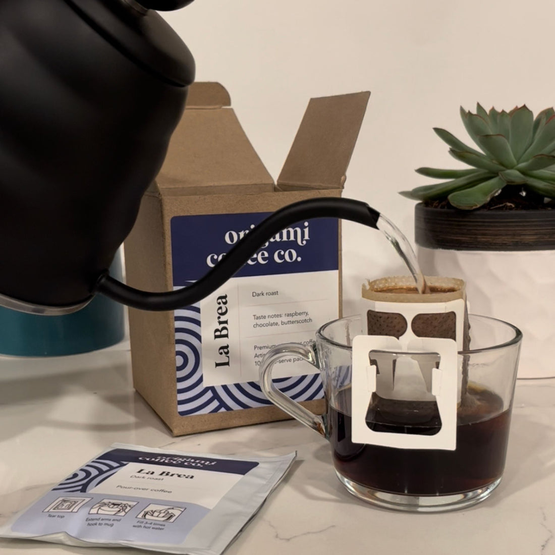 Origami Coffee Co. single-serve pour-over coffee pack in a glass mug with a hario buono gooseneck kettle and La Brea Dark Roast 10-pack box