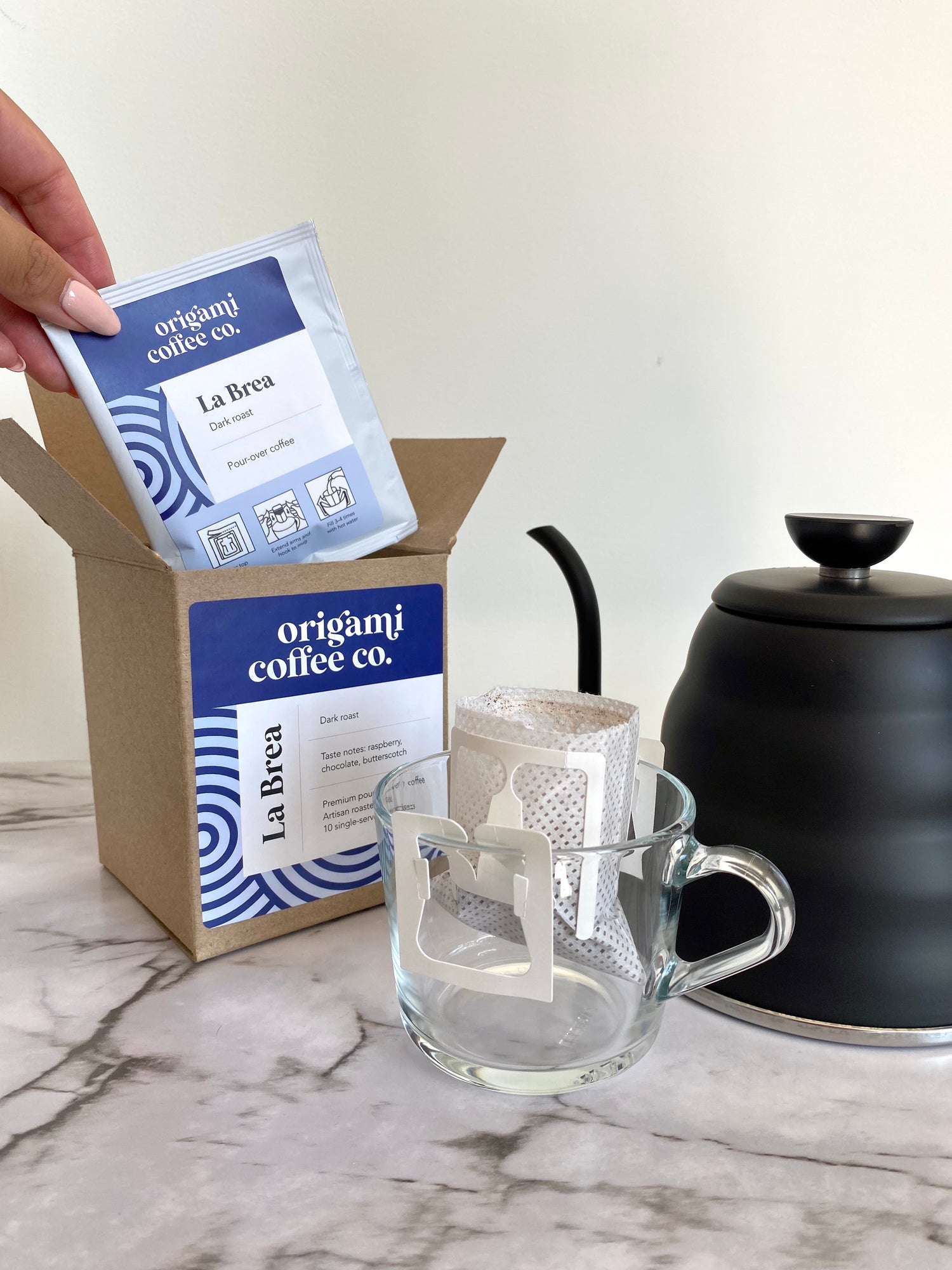 Origami Coffee Co. single-serve pour-over coffee pack with box, glass mug, and matte black Hario kettle.